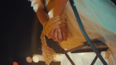 Bir insanın ellerinin iple bağlandığı, sandalyeye oturduğu yakın çekim. Şahıs akıcı beyaz bir elbise giyiyor, ve arka plan ışıklarla hafifçe aydınlatılıyor, dramatik bir atmosfer yaratıyor..