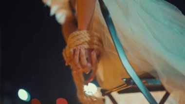 Sandalyeye oturmuş, akıcı beyaz bir kıyafet giymiş halat tutan birinin yakın çekimi. Arka planda bulanık ışıklar var, rüya gibi bir atmosfer yaratıyor..