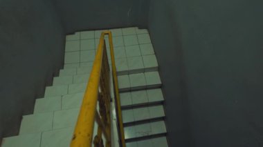 Merdivenin tepesinden beyaz kiremitli merdivenler ve sarı korkulukların olduğu bir manzara. Duvarlar sessiz bir renge boyanmış, basit ve işlevsel bir atmosfer yaratıyor..