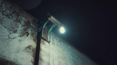 Geceleyin loş bir ara sokak, yıpranmış duvarlar ve manzarayı aydınlatan bir sokak lambası. Atmosfer sessiz ve tüyler ürpertici. Işığın yansıttığı gölgeler var. Uzakta birkaç park edilmiş araç görülüyor, konutlarla çevrili..