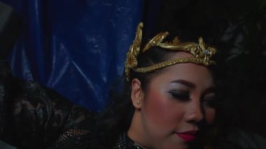 Dramatik makyajlı ve altın bir saç bandı takmış uzanmış gülümseyen bir kadın. Siyah dantel bir elbise giyiyor..