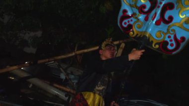 Geleneksel giyinmiş bir insan elinde uzun bir nesne, muhtemelen bir mızrak, arka planda yeşillik olan karanlık bir ortamda..