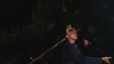 Geleneksel kıyafetler ve altın başlık takan bir insan, gece kültürel bir dans sergiliyor. Arka plan koyu renkli ve bazı yapraklar görülebilir..