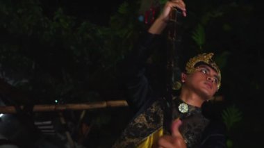 Geleneksel giysiler içinde bir kişi gece bir ayin ya da merasim gerçekleştiriyor, bambu yapılarıyla çevrili..