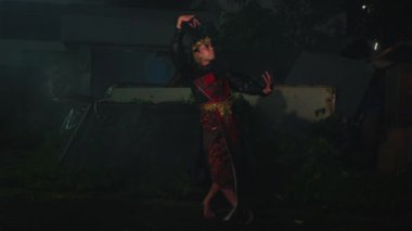Geleneksel kıyafetli biri gece dans ediyor. Kostüm kırmızı ve siyah renklerle özenle hazırlanmış ve dansçı anlamlı el hareketleri yapıyor..