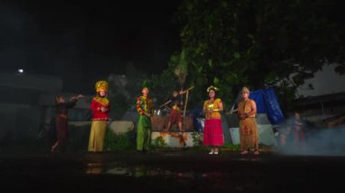 Geleneksel kostümlü ve makyajlı bir grup sanatçı gece dışarıda kültürel bir dans veya tiyatro gösterisi düzenliyor. Arkaplanda ağaçlar ve bir bina var.