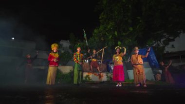 Bir grup geleneksel kostümlü sanatçı gece açık hava sahnesinde. Arka planda ağaçlar ve atmosferi güçlendiren sis veya duman efektleri bulunan bir bina yer alıyor..