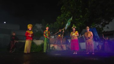 Geleneksel kostümler giymiş bir grup insan gece kültürel dans gösterisi yapıyor. Arkaplanda ağaçlar ve binalar var.