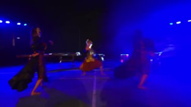 Mavi sahne ışıkları altında dans eden üç kişinin bulanık görüntüsü.