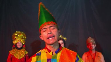 Renkli geleneksel bir kostüm ve uzun yeşil bir şapka giyen bir kişi, arka planda iki sanatçıyla birlikte sahnede performans sergiliyor..