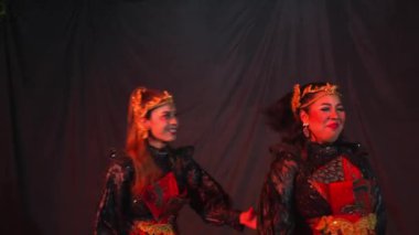 Geleneksel, süslenmiş, altın başlıklı iki kadın koyu bir arkaplanda dikiliyorlar. Bir kadın gülümserken diğeri tarafsız bir ifade takınıyor..