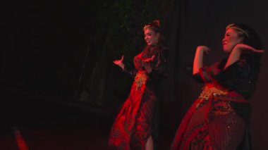Geleneksel giyinmiş iki kadın, kırmızı ışıkla karanlık bir sahnede kültürel dans ediyorlar..
