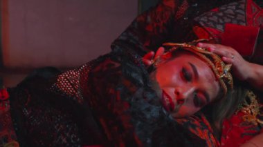 Geleneksel giyimli, karmaşık başlıklı, uzanmış ve duygusal görünen biri. Sahne kırmızı ışıkla aydınlatılmış, dramatik bir atmosfer yaratıyor..