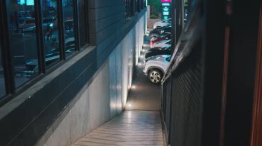 Otoparka inen loş bir merdiven. Duvarlar şehir ışıklarını yansıtan pencerelerle kaplı. Otomobiller park halinde, sokak lambaları ve yakındaki tabelalarla aydınlatılıyor, geceleri canlı bir şehir atmosferi yaratıyor..