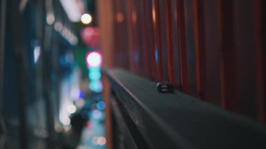 Modern bir açık hava barında ya da restoranda canlı bir gece sahnesi. Renkli neon ışıklar uzayı aydınlatarak canlı bir atmosfer yaratıyor. Bir grup insan masalara oturmuş, içki ve sohbetin tadını çıkarıyor, şehrin ışıklarıyla. 