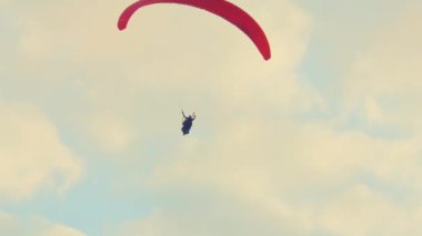 Bulutlu bir gökyüzüne karşı kırmızı bir paraşütle yürüyen biri. Sahne maceranın heyecanını ve açık hava sporlarının güzelliğini yakalıyor..