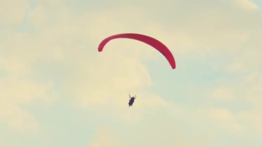 Yumuşak, bulutlu bir gökyüzünde süzülen yalnız bir paraglider. Paraşütçü, soluk mavi ve beyaz bulutlara karşı parlak kırmızı bir gölgeliğe sahiptir. Sahne maceranın heyecanını ve hava sporlarının güzelliğini yakalıyor..
