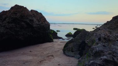 Her iki taraftaki kayalık oluşumların durgun bir denize yol açtığı sakin bir sahil manzarası. Güneş batarken gökyüzü yumuşak pastel renklerle boyanır ve suyun yüzeyine yansıtır. Kumda küçük kayalar ve yeşil yosun parçaları görülebilir..