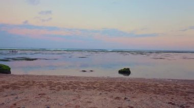 Sahilde büyük kayalık oluşumların olduğu sakin bir sahil sahnesi ve arka planda sakin bir deniz. Güneş batarken gökyüzü yumuşak pastel renklerle boyanır ve sakin bir atmosfer yaratır.. 