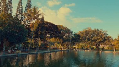Bereketli yeşillikler ve uzun ağaçlarla çevrili sakin bir göletin olduğu sakin bir manzara. Gökyüzü birkaç bulutla temiz, barışçıl bir atmosfer yaratıyor..