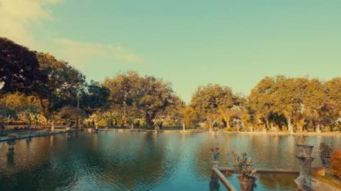 Bereketli yeşillik ve ağaçlarla çevrili sakin bir göletin olduğu sakin bir manzara. Gökyüzü bulutların zerresiyle berrak, barışçıl bir atmosfer yaratıyor..