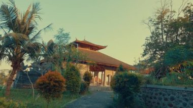Yeşillik ve palmiye ağaçlarıyla çevrili geleneksel bir Asya tapınağı manzarası. Girişe giden bir patika var. Sıcak güneş ışığı sahnenin üzerine altın bir renk saçar ve huzurlu bir atmosfer yaratır..