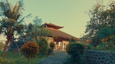 Yeşillik ve palmiye ağaçlarıyla çevrili sazdan çatılı geleneksel bir binanın bulunduğu sakin bir manzara. Yol girişe çıkıyor, sıcak güneş ışığıyla yıkanmış..