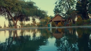 Sakin bir gölet, ağaçları ve sazdan çatılı bir kulübeyi yansıtan sakin bir manzara. Yeşillik suları sarar ve huzurlu bir atmosfer yaratır..