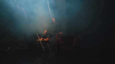 Bir sanatçı, dumanla çevrili loş bir alanda bir sandalyeye oturur. Atmosfer dramatik, yakınlarda ahşap bir masa var. Sahne bir gizem ve içgözlem hissi veriyor..