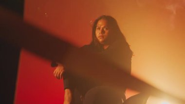 Düşünceli bir kadın sıcak, turuncu bir arka planda oturuyor, yumuşak ışıklandırma karamsar bir atmosfer yaratıyor. Derin düşüncelere dalmış, rahat bir duruşu var..