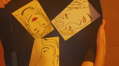 Sarı kağıt üzerinde üç sanatsal yüz çizimi, abartılı özellikleri olan basit çizgi çizimleri. Çizimler yansıtıcı bir yüzeye yerleştiriliyor ve ilginç bir görsel etki yaratıyor..