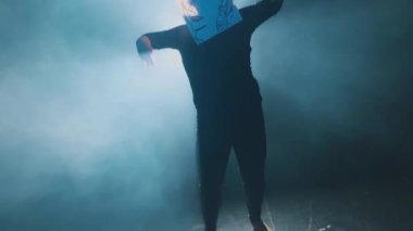 Karanlık bir ortamda yüzünü kaplayan bir çizimle poz veren biri, dumanla çevrilidir. Eserde, basit çizgileri ve sakin bir ifadesi olan stilize bir yüz yer alıyor..