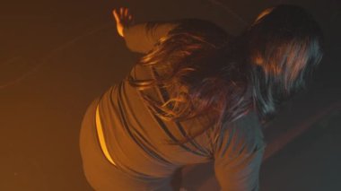 Uzun saçlı bir insan, arkadan bakıldığında, tek koluyla uzanır. Sahne sıcak ışıkla aydınlatılıyor ve dramatik bir atmosfer yaratıyor. Arkaplan koyu renkli, şekle odaklanmayı güçlendiriyor.