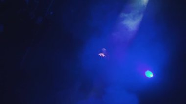Sahnede mavi ve mor ışık efektleriyle çevrili bir sanatçı, duman ve gölgelerle kasvetli bir atmosfer yaratıyor..