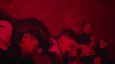 Kırmızı ışıklandırmalı loş bir ortamda üç kişinin olduğu dramatik bir sahne. Siyah ve beyaz çizgili kıyafetler giyiyorlar, çarpıcı bir görsel karşıtlık yaratıyorlar.. 