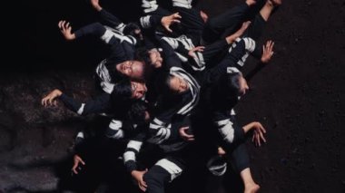 Siyahi kıyafetli genç bir adamın, parlak bir spot ışıklarıyla aydınlanan, kendinden emin bir şekilde ön planda durduğu dramatik bir sahne. Arka planda, başka bir figür sahne malzemesi silahı tutuyor, bu da gerilimi gösteriyor.. 