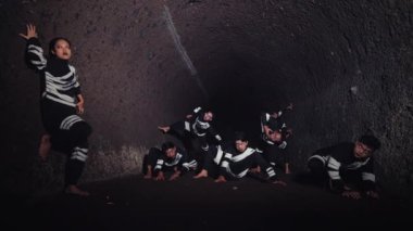 Siyahi kıyafetli genç bir adamın, parlak bir spot ışıklarıyla aydınlanan, kendinden emin bir şekilde ön planda durduğu dramatik bir sahne. Arka planda, başka bir figür sahne malzemesi silahı tutuyor, bu da gerilimi gösteriyor.. 