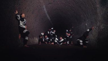 Siyahi kıyafetli genç bir adamın, parlak bir spot ışıklarıyla aydınlanan, kendinden emin bir şekilde ön planda durduğu dramatik bir sahne. Arka planda, başka bir figür sahne malzemesi silahı tutuyor, bu da gerilimi gösteriyor.. 