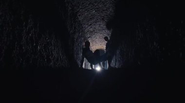 Karanlık bir tünelin sonunda duran siluetli figürler, parlak bir ışıkla aydınlatılıyor. Tünel duvarları pürüzlü ve dokulu, gizemli bir atmosfer yaratıyor..