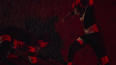 Koyu renk elbiseli iki kişi kırmızı zemine karşı dinamik bir duruş sergiliyor. Bu da hareket ve yoğunluğu gösteriyor. Sahne gölgeler ve röflelerle dramatik.