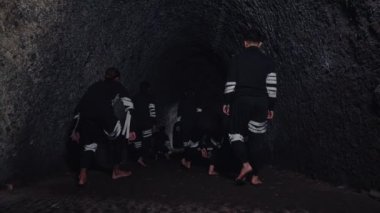 Siyahi kıyafetli genç bir adamın, parlak bir spot ışıklarıyla aydınlanan, kendinden emin bir şekilde ön planda durduğu dramatik bir sahne. Arka planda, başka bir figür sahne malzemesi silahı tutuyor, bu da gerilimi gösteriyor.. 