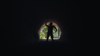 Karanlık bir tünelde, kafasının üstünde bir nesneyi tutan bir kişinin silüeti. Arka plan tünelin dışındaki yeşilliği ortaya çıkarıyor, ışık ve gölge arasında bir kontrastlık oluşturuyor..