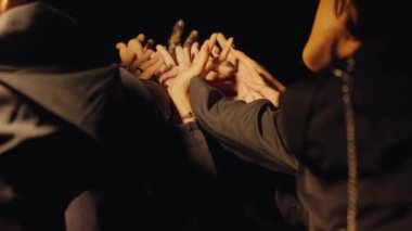 Siyahi kıyafetli genç bir adamın, parlak bir spot ışıklarıyla aydınlanan, kendinden emin bir şekilde ön planda durduğu dramatik bir sahne. Arka planda, başka bir figür sahne malzemesi silahı tutuyor, bu da gerilimi gösteriyor.. 