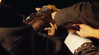 Siyahi kıyafetli genç bir adamın, parlak bir spot ışıklarıyla aydınlanan, kendinden emin bir şekilde ön planda durduğu dramatik bir sahne. Arka planda, başka bir figür sahne malzemesi silahı tutuyor, bu da gerilimi gösteriyor.. 