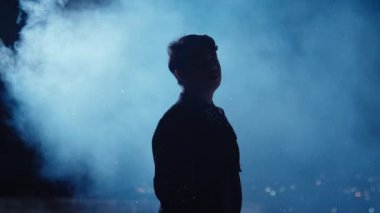 Dumanlı mavi arka planda duran bir kişinin silueti dramatik bir atmosfer yaratıyor. Bu figür kısmen aydınlatılmış ve gizemli titreşime eklenmiş..