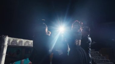 Bir grup şık giyimli kişi arka planda uyarı bandıyla loş bir ortamda poz veriyorlar. Esas konu, dekoratif unsurları olan siyah kıyafetli genç bir adamın doğrudan kameraya bakması..