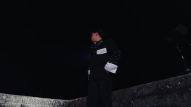 Gece bir çıkıntıda duran, beyaz kelepçeli, şık siyah bir kıyafet giyen biri. Sahne dramatik, karanlık bir çevre ve figürü vurgulayan bir spot ışığı etkisi var..