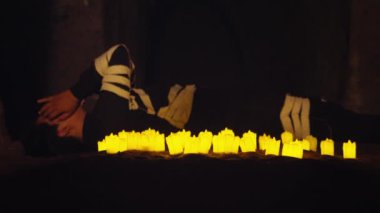 Karanlık bir ortamda, sarı mumlarla çevrili yerde yatan biri mistik bir atmosfer yaratıyor..