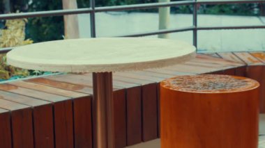 Yuvarlak taş bir masa ve silindirik turuncu bir tabureye sahip modern bir açık hava oturma alanı. Masanın su damlacıkları ile pürüzsüz bir yüzeyi var ve arka planda ahşap döşeme ve yeşillik içeriyor..