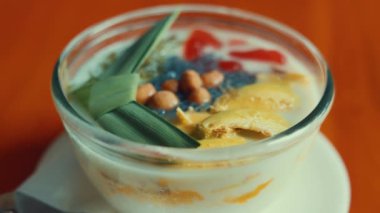Cam bir kasede, hindistan cevizi sütü, meyve ve kırmızı jöle ve yeşil pandan yaprakları gibi renkli malzemeler içeren bir tatlı..